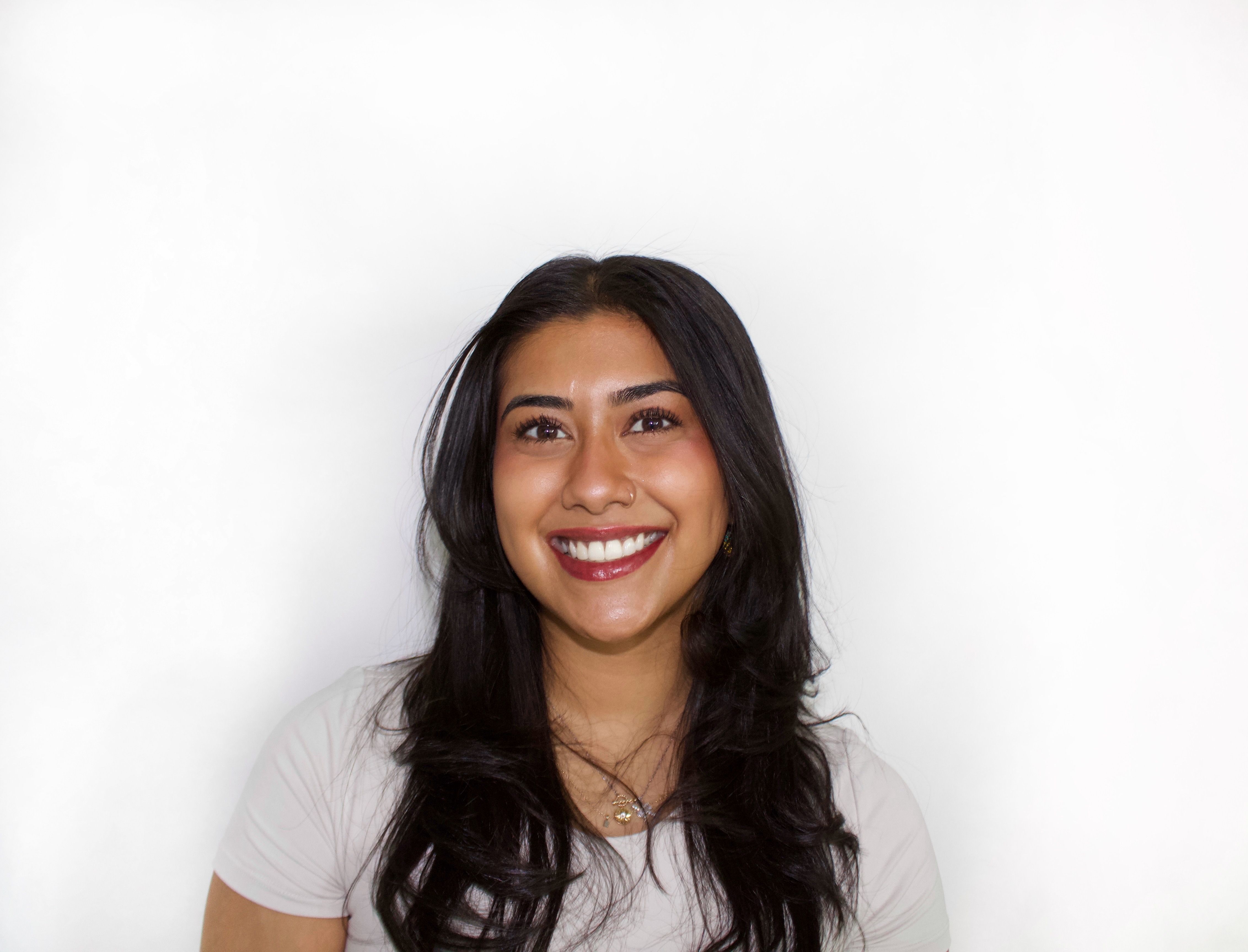 A woman with long black hair, standing against a white background and smiling brightly, showing her teeth