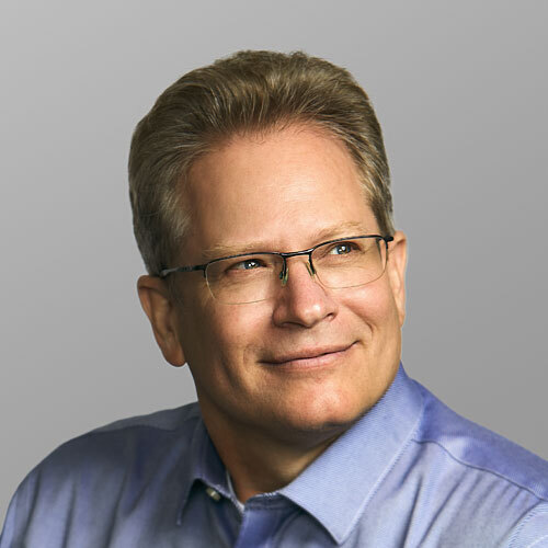 A man in a blue dress shirt and glasses, smiling while looking off to the side