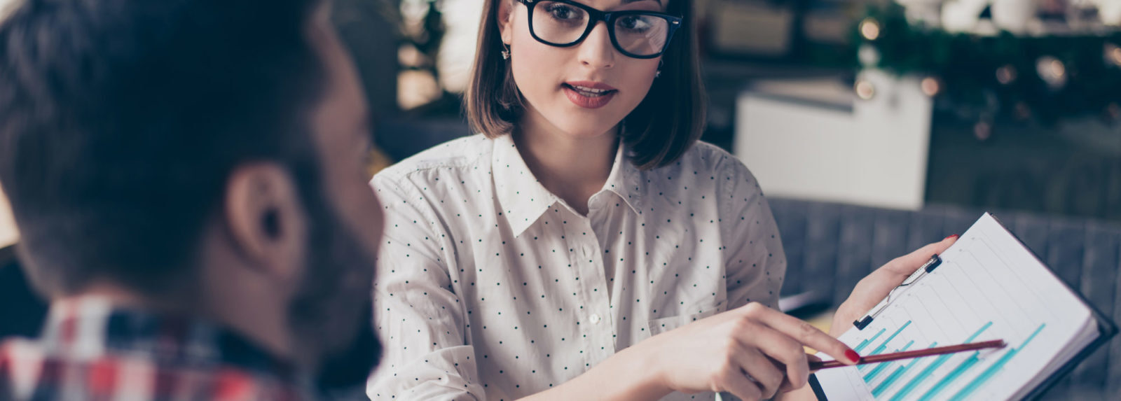 A woman wearing glasses, holding a paper, and gesturing to it with her pen while speaking to her colleague
