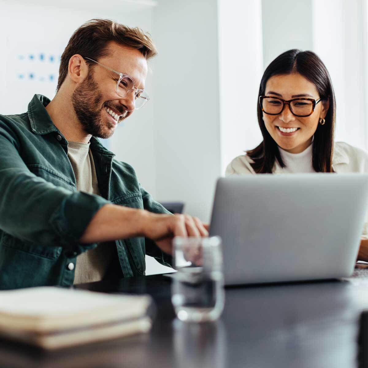 A man and a woman, both wearing glasses, sit together smiling at their laptop screen